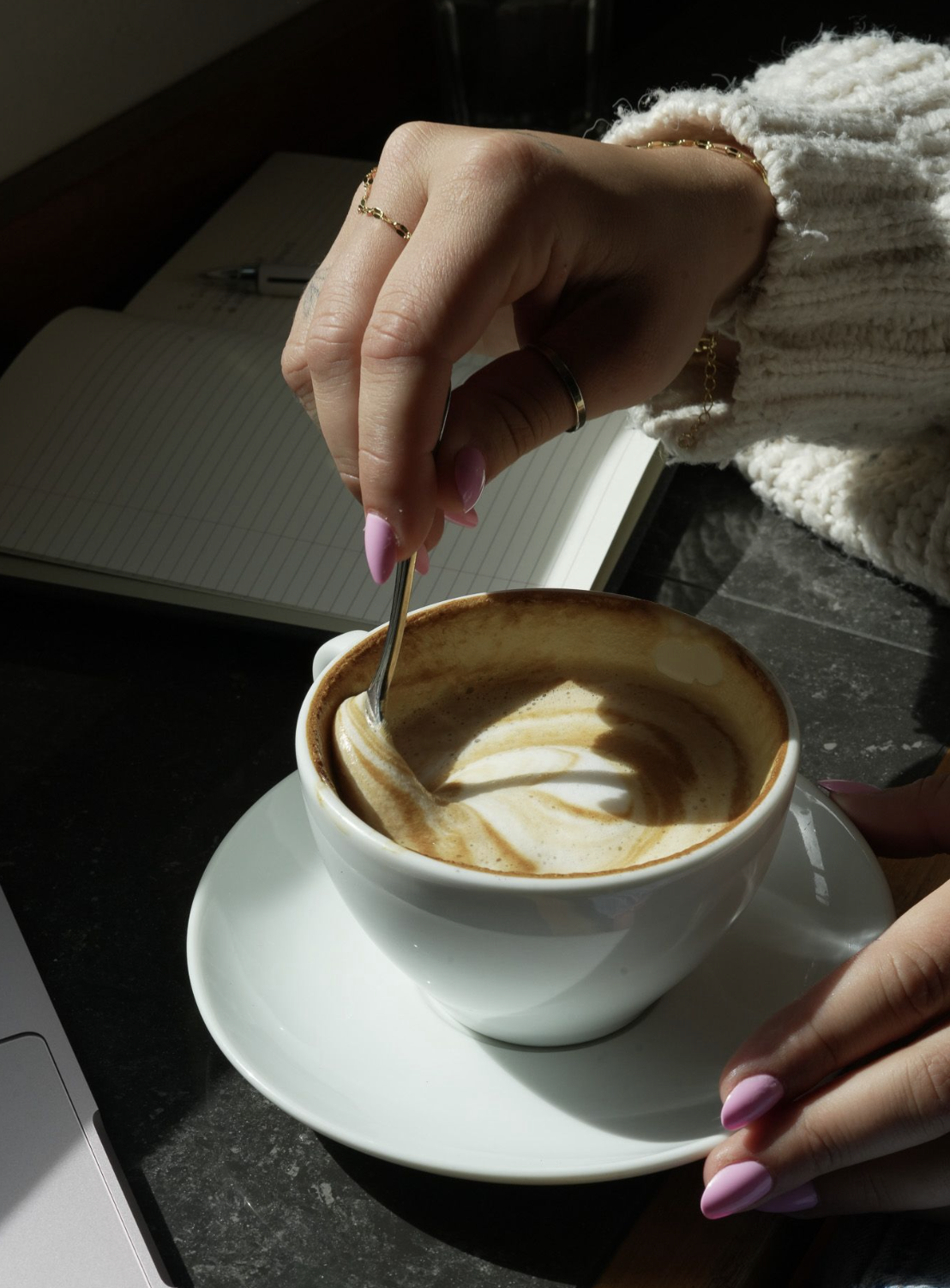 A hand stirring a cappuccino beside an open notebook